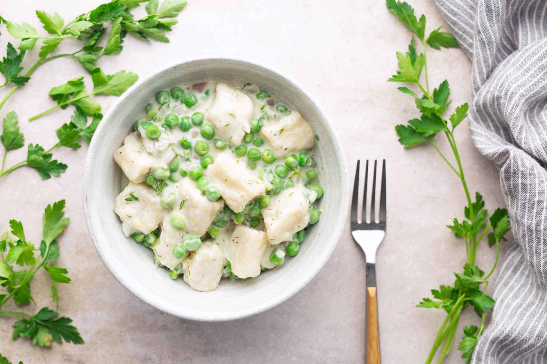 A bowl of gnocchi in a creamy sauce with green peas, surrounded by fresh parsley sprigs and a fork on the side.