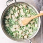 A skillet filled with potato gnocchi and green peas in a creamy sauce, being stirred with a wooden spoon.
