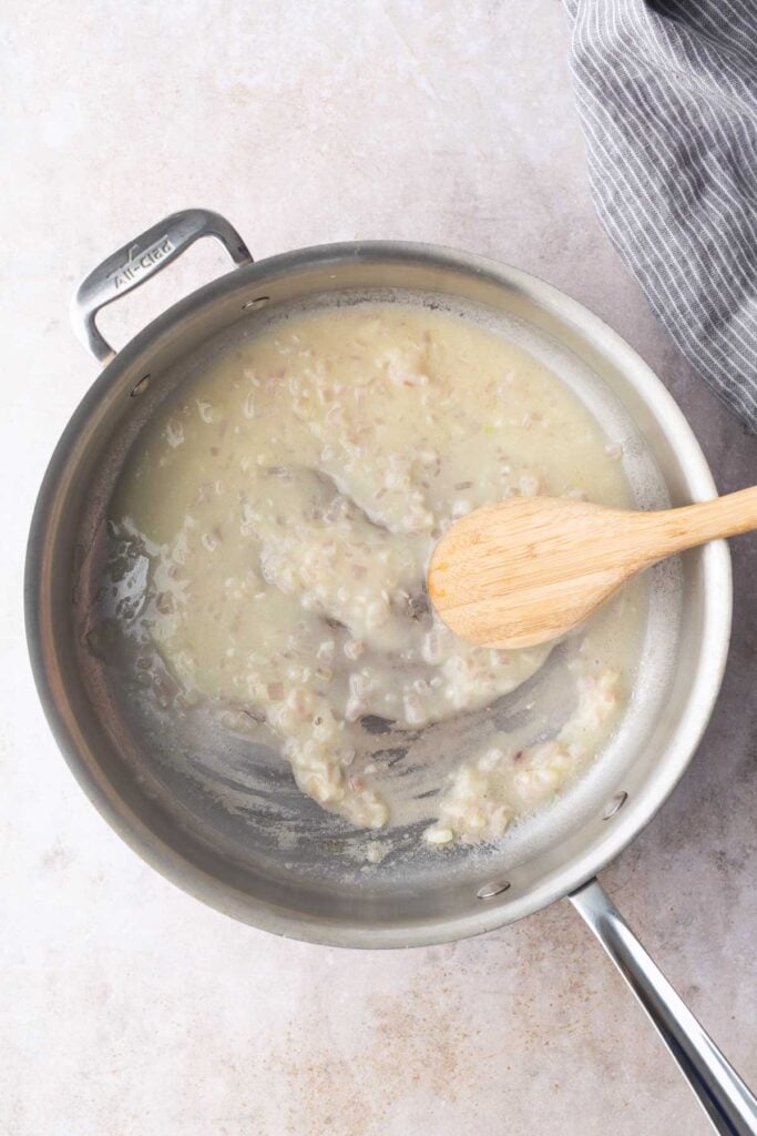 A metal pan with a creamy mixture being stirred by a wooden spoon on a light countertop, with a striped cloth nearby.