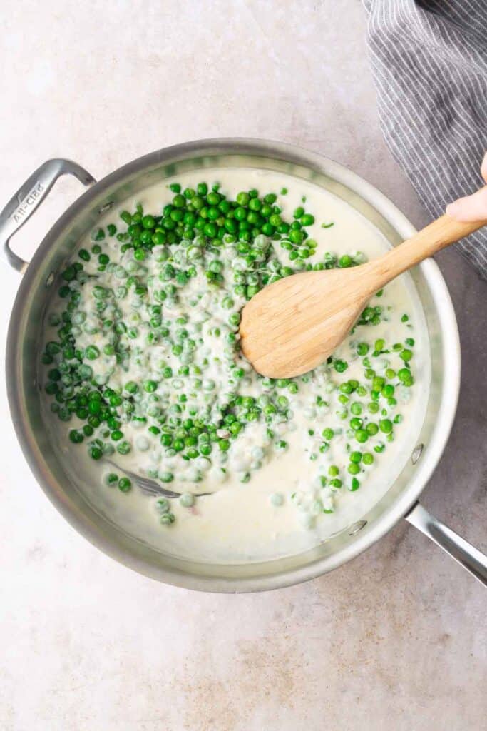 A saucepan filled with creamy sauce and green peas being stirred with a wooden spoon on a light countertop.