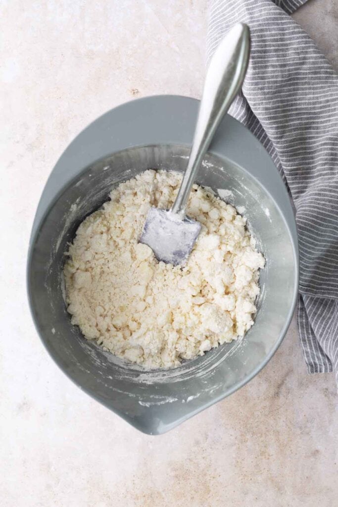 A mixing bowl with coarse, crumbly dough and a spatula, next to a gray-and-white striped cloth on a light countertop.