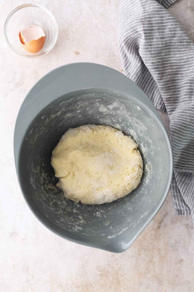 A gray mixing bowl with dough inside, next to a cracked egg in a glass bowl and a gray-striped kitchen towel on a light countertop.