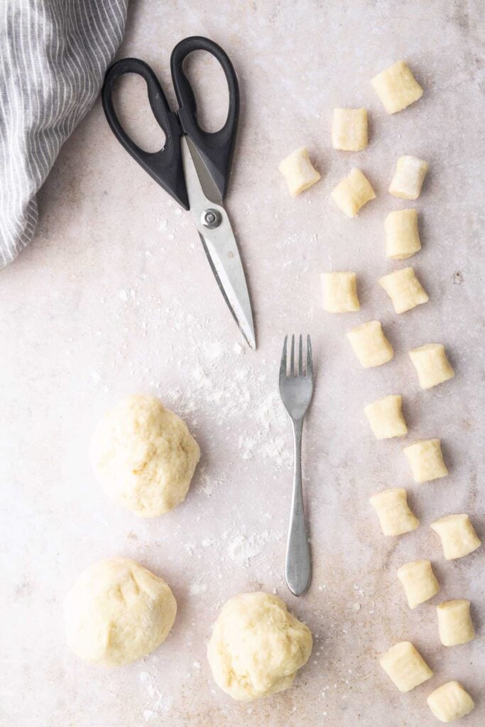 Balls of dough, dough pieces cut into small chunks, a fork, and a pair of kitchen scissors are arranged on a floured surface with a striped cloth in the corner.