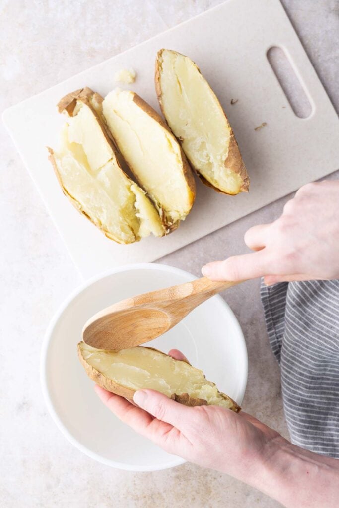 A person scoops the inside of a baked potato half with a wooden spoon over a white bowl; other potato halves are on a cutting board.