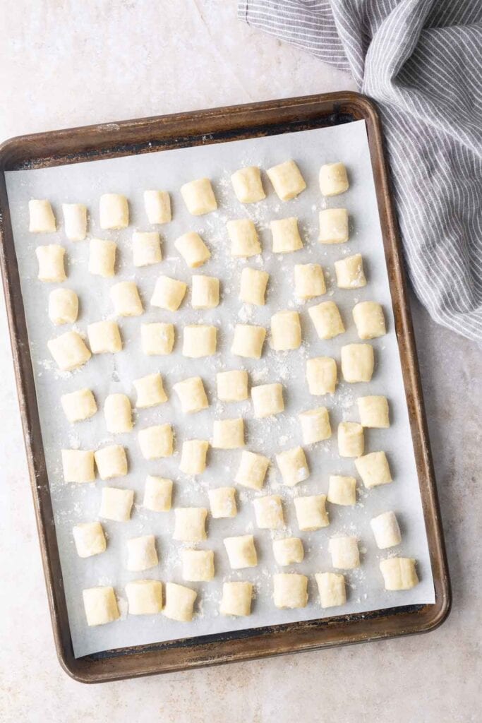 A baking sheet lined with parchment paper holds evenly spaced, uncooked gnocchi pieces. A gray striped cloth is partially visible in the top right corner.