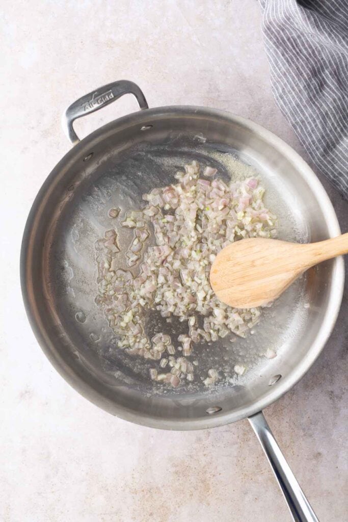 Chopped shallots are being sautéed in a stainless steel pan with a wooden spoon on a light countertop.