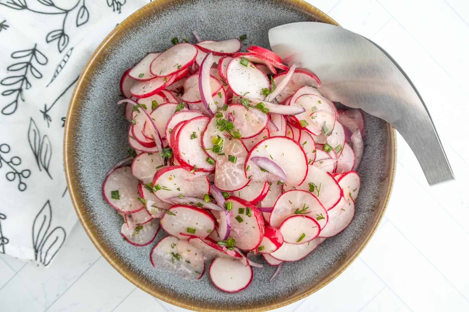 A bowl of sliced radish and red onion salad garnished with chopped herbs, with a serving utensil on the side.