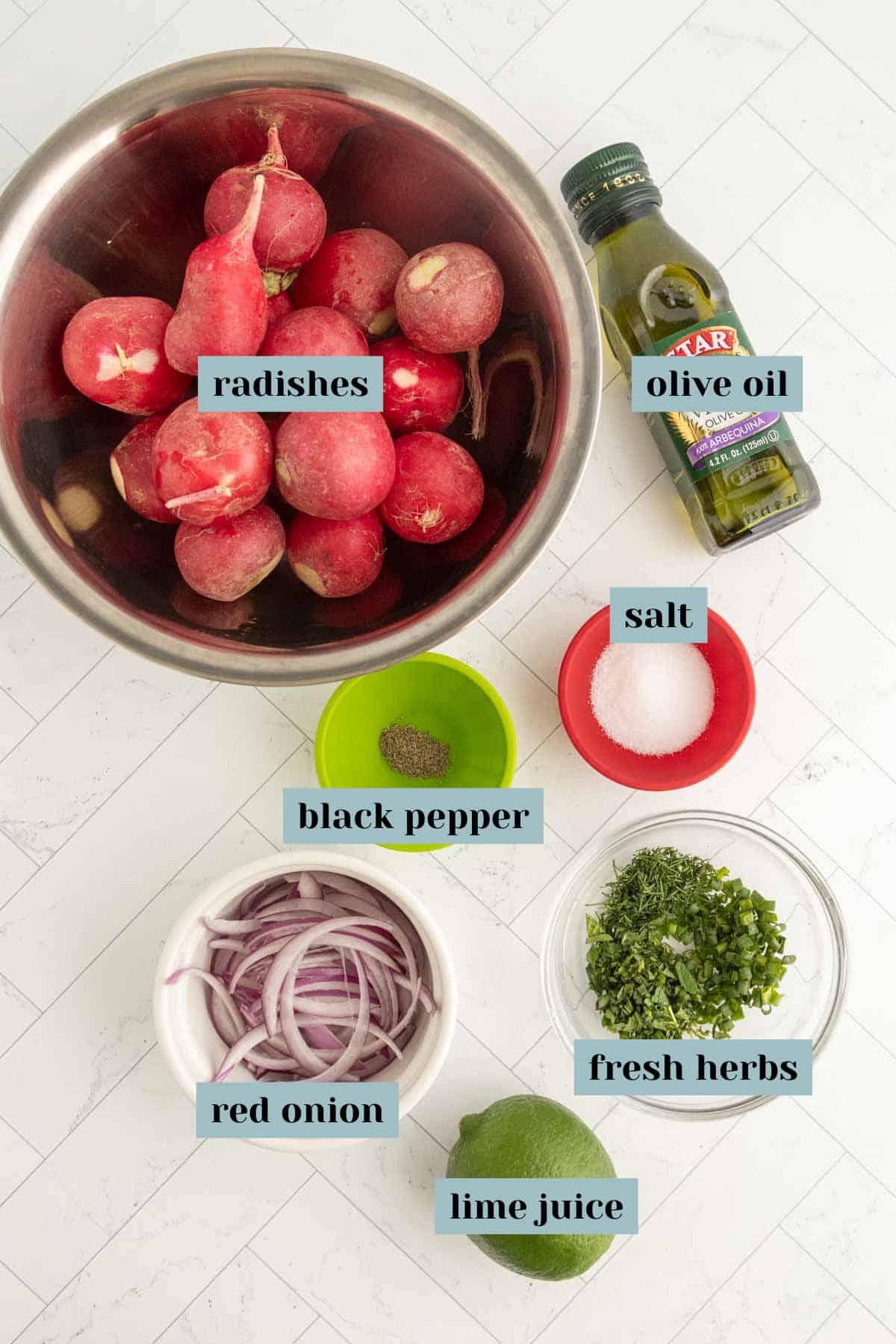 A bowl of whole radishes, a bottle of olive oil, salt, black pepper, sliced red onion, chopped fresh herbs, and a halved lime arranged on a white surface.