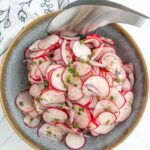 A bowl of sliced radishes and red onions garnished with herbs, with metal tongs resting on the edge of the bowl.