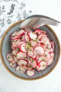 A bowl of sliced radishes and red onions garnished with herbs, with metal tongs resting on the edge of the bowl.