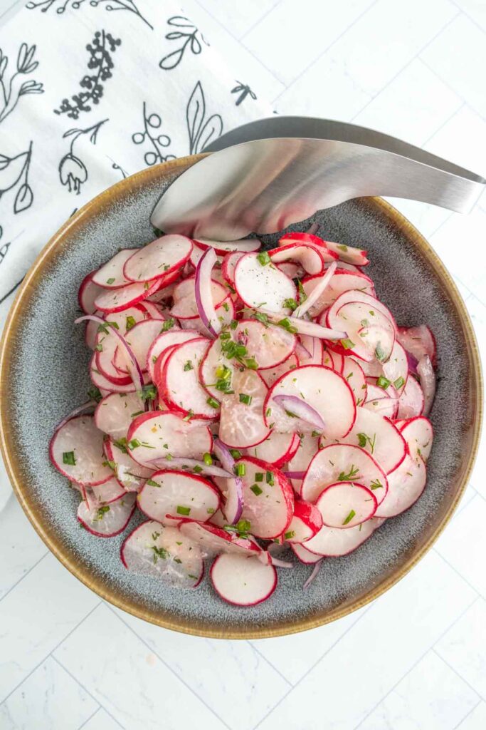 A bowl of sliced radishes and red onions garnished with herbs, with metal tongs resting on the edge of the bowl.
