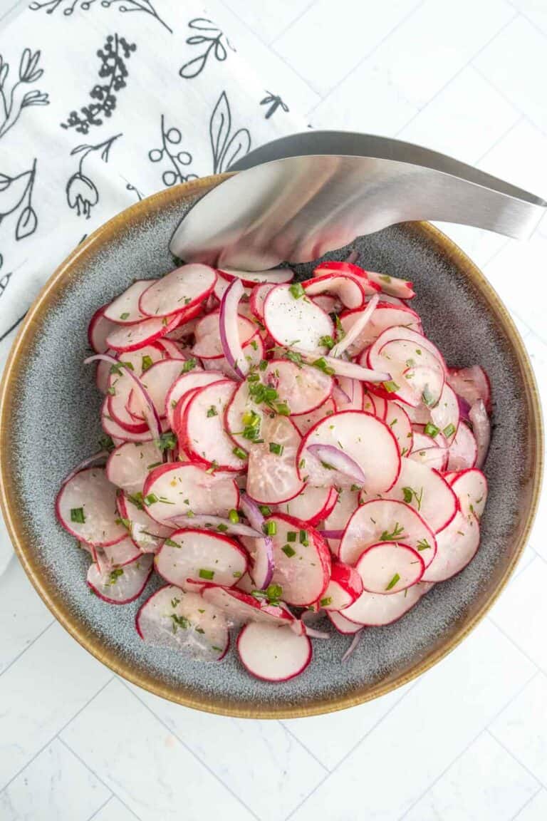 A bowl of sliced radishes and red onions garnished with herbs, with metal tongs resting on the edge of the bowl.