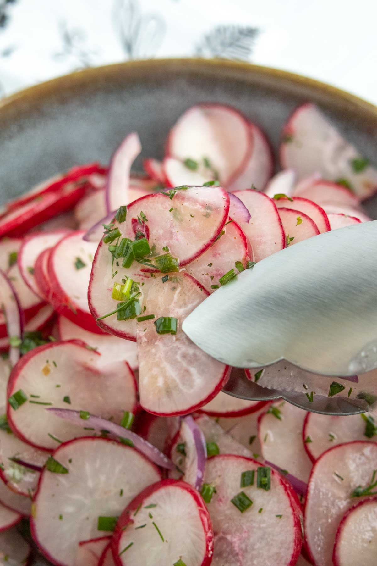A close-up of metal tongs holding thinly sliced radishes garnished with chopped herbs, over a bowl filled with more sliced radishes and red onions.