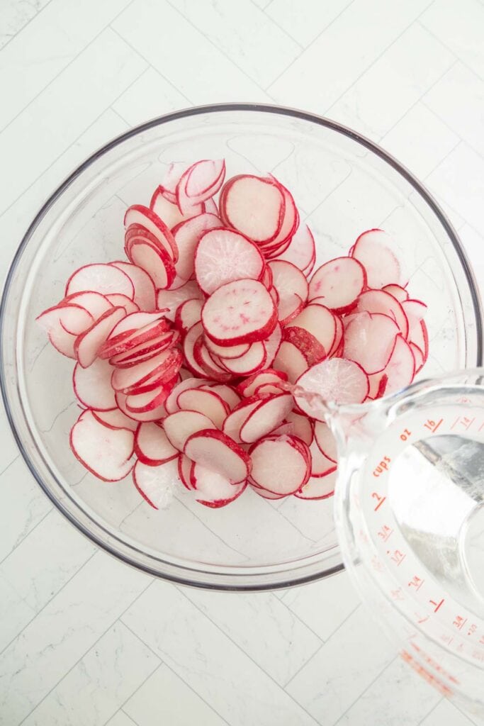 A glass bowl filled with thinly sliced radishes is shown, with a measuring cup of water being poured into the bowl on a white tiled surface.
