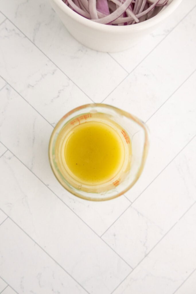 A glass measuring cup with yellow liquid on a white tiled surface, next to a bowl containing sliced red onions.