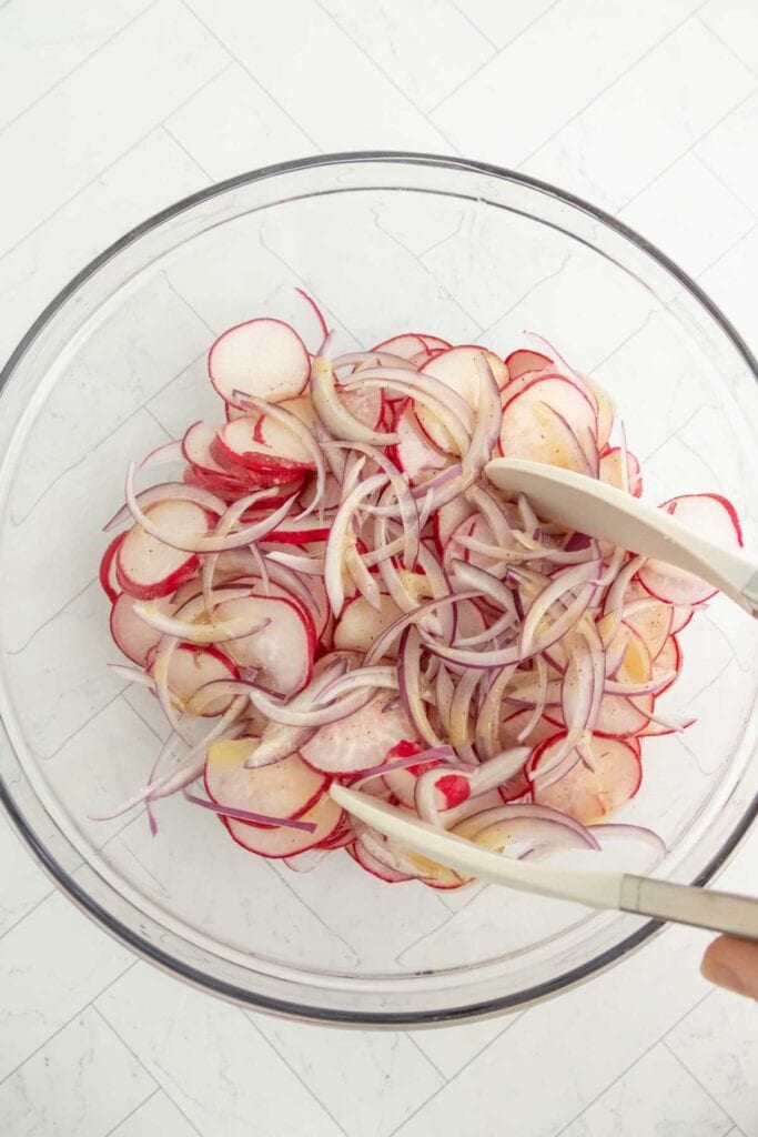 A glass bowl containing sliced radishes and red onions being mixed with tongs on a white countertop.