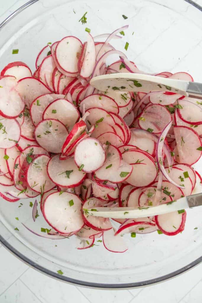 Thinly sliced radishes and red onions mixed with chopped herbs in a glass bowl, with a pair of white tongs.