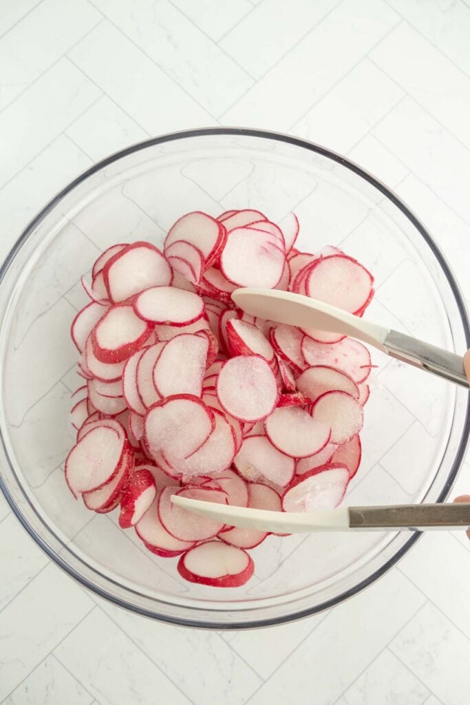 A clear glass bowl filled with thinly sliced radishes, being mixed with white kitchen tongs on a white tiled surface.