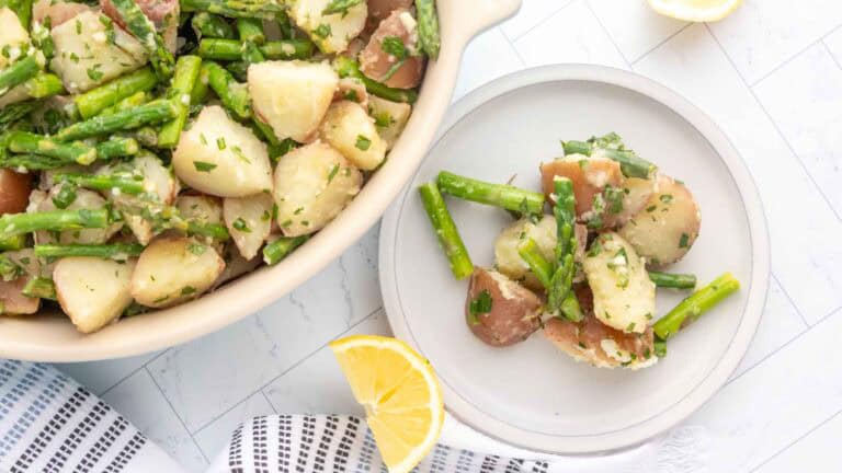 A bowl and plate of potato and asparagus salad garnished with herbs, next to lemon wedges on a white tiled surface.