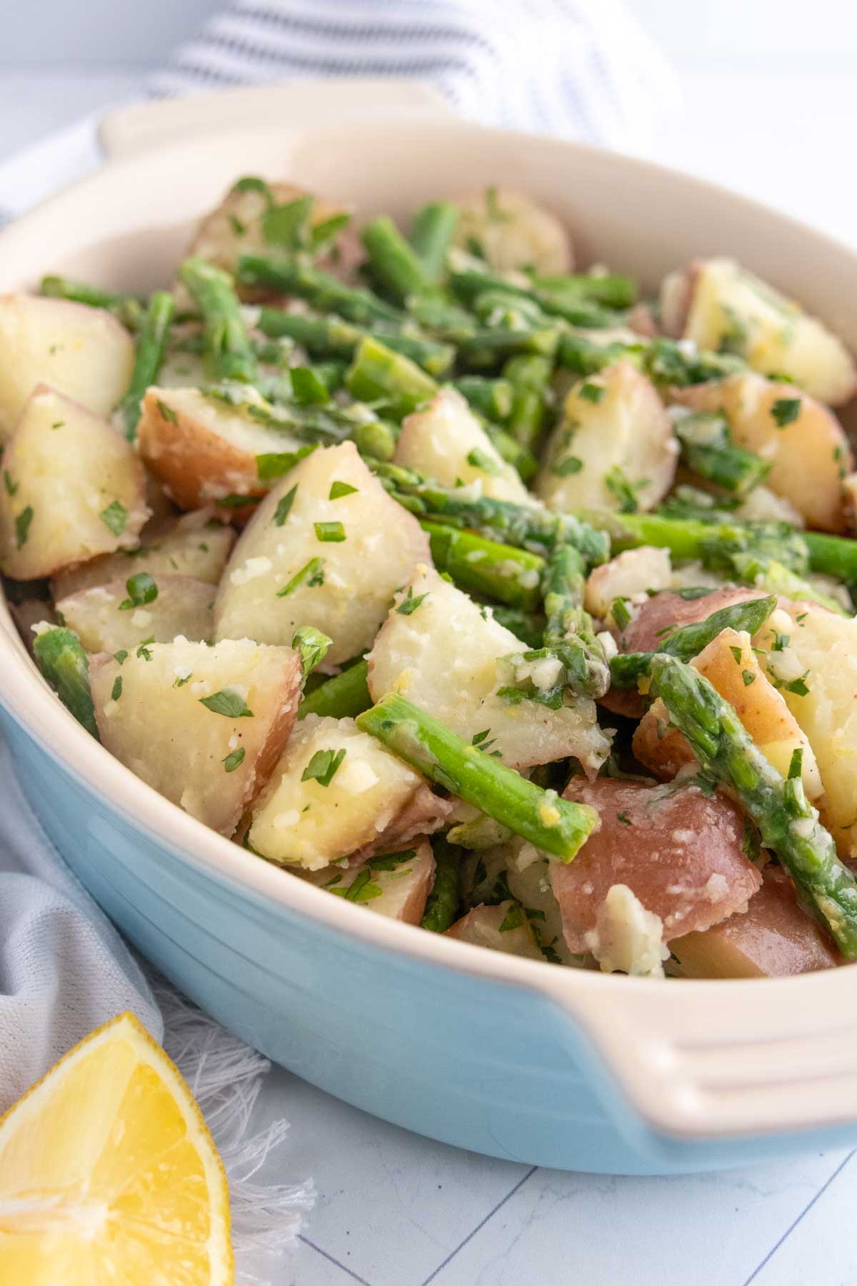 A dish filled with cooked red potatoes and asparagus spears, garnished with chopped herbs, sits on a table next to a lemon wedge.