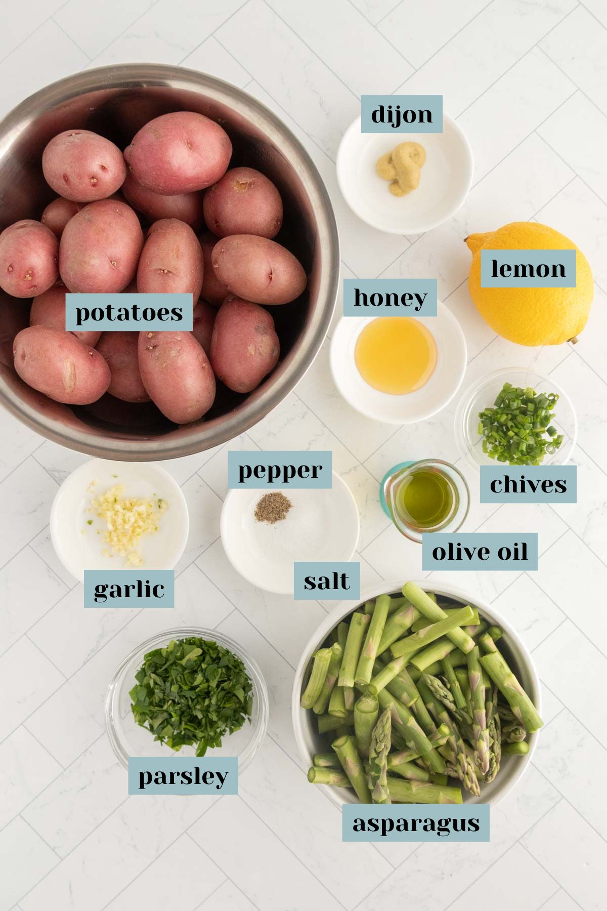 Overhead view of raw potatoes, asparagus, chopped parsley, chives, garlic, salt, pepper, olive oil, honey, dijon mustard, and a lemon arranged in bowls on a white surface.