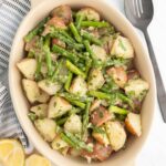 A ceramic dish filled with a salad of chopped red potatoes and green beans, garnished with herbs, next to a slotted spoon, linen napkin, and lemon wedges.