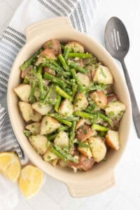 A ceramic dish filled with a salad of chopped red potatoes and green beans, garnished with herbs, next to a slotted spoon, linen napkin, and lemon wedges.