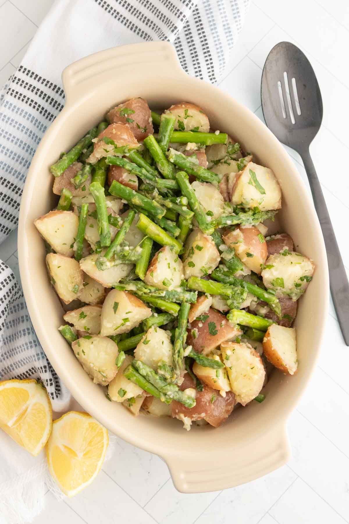 A ceramic dish filled with a salad of chopped red potatoes and green beans, garnished with herbs, next to a slotted spoon, linen napkin, and lemon wedges.