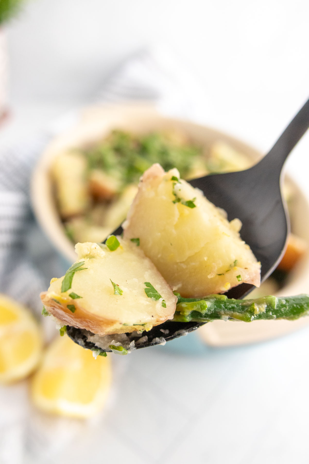 A black spoon holds pieces of cooked potato garnished with chopped parsley, with a dish of similar potatoes and lemon wedges in the background.