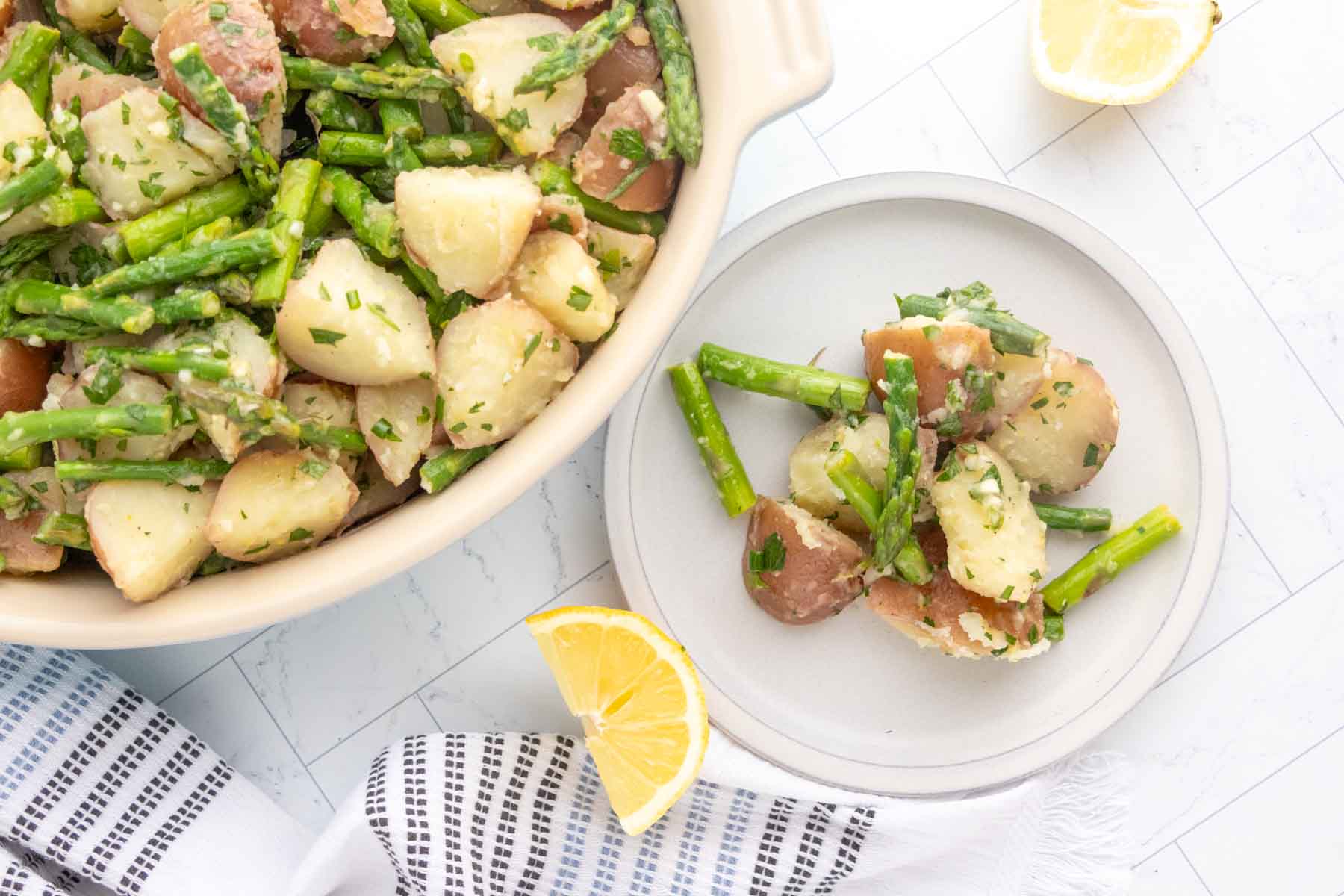 Bowl and plate of potato and asparagus salad garnished with herbs, next to lemon wedges on a white surface with a striped cloth.