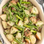 Oval dish filled with warm potato salad made of red potatoes and asparagus, garnished with herbs. Text overlay reads "Warm Potato Salad.