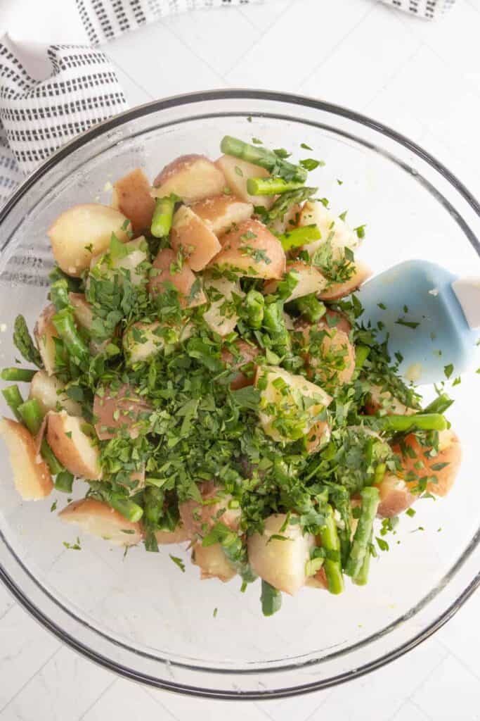 A glass bowl filled with a potato salad made of chopped red potatoes, asparagus, and fresh herbs, with a blue spatula on the side.