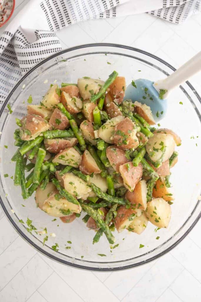 A glass bowl filled with a salad of chopped red potatoes, green beans, and parsley, being mixed with a blue spatula.