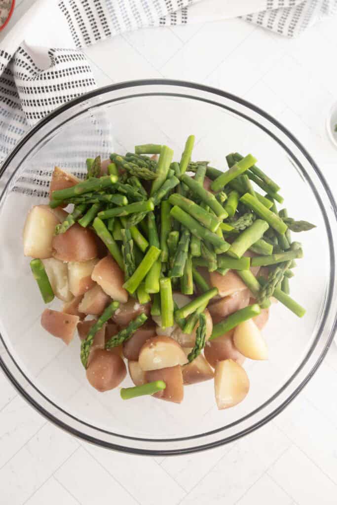 A glass bowl filled with chopped red potatoes and asparagus pieces, placed on a white countertop next to a striped kitchen towel.