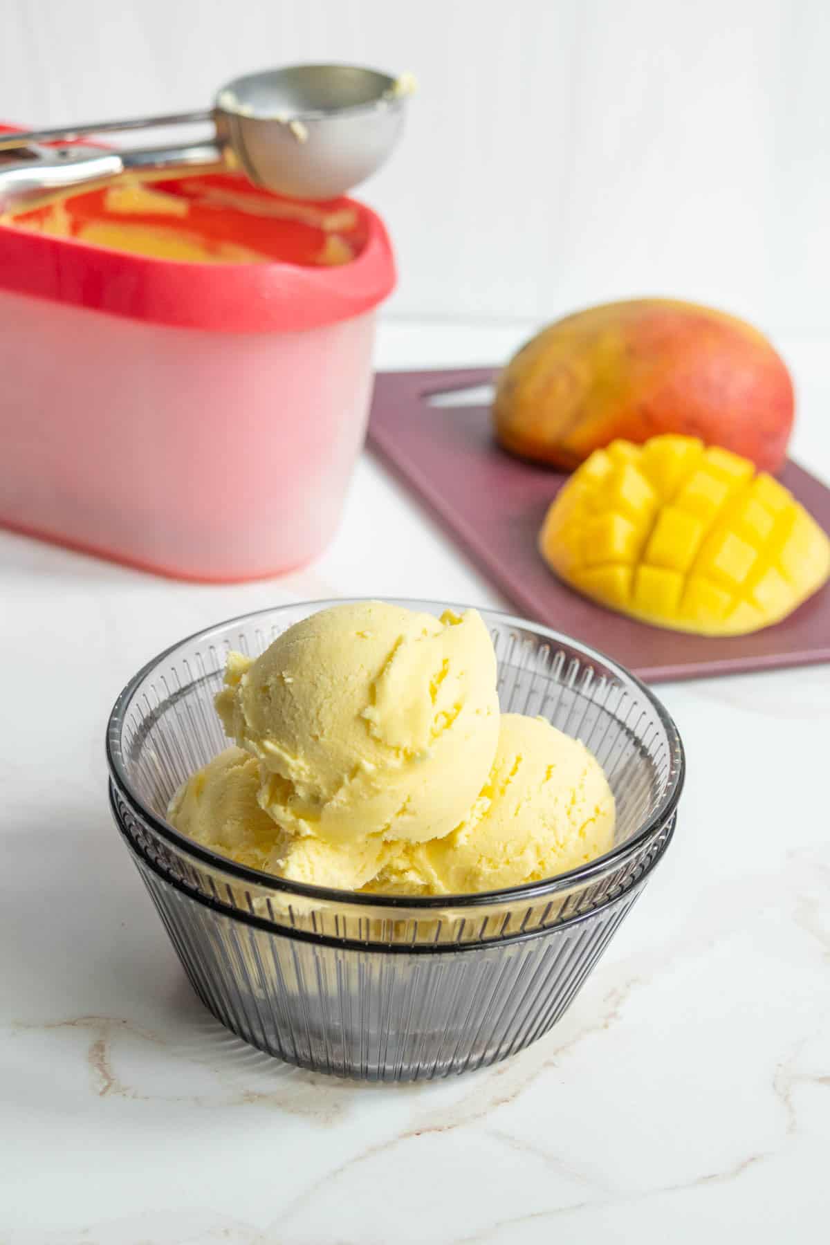 A glass bowl with two scoops of mango ice cream sits on a counter; behind it are a sliced mango, a whole mango, an ice cream container, and a scoop.