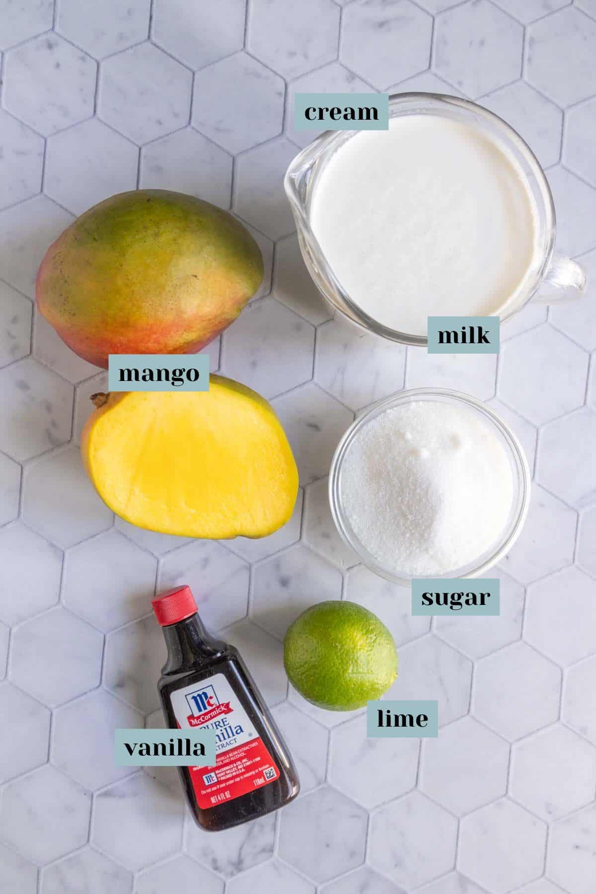 Ingredients on a hexagonal tile surface: whole mango, half mango, lime, bowl of sugar, glass measuring cup of milk, glass measuring cup of cream, bottle of vanilla extract. Labels identify each item.