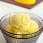 A glass bowl filled with three scoops of yellow mango ice cream, with fresh mango pieces on a cutting board in the background.