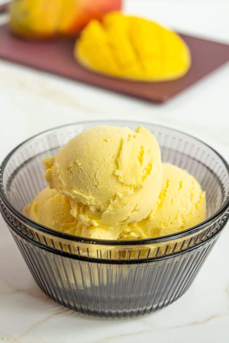 A glass bowl filled with three scoops of yellow mango ice cream, with fresh mango pieces on a cutting board in the background.