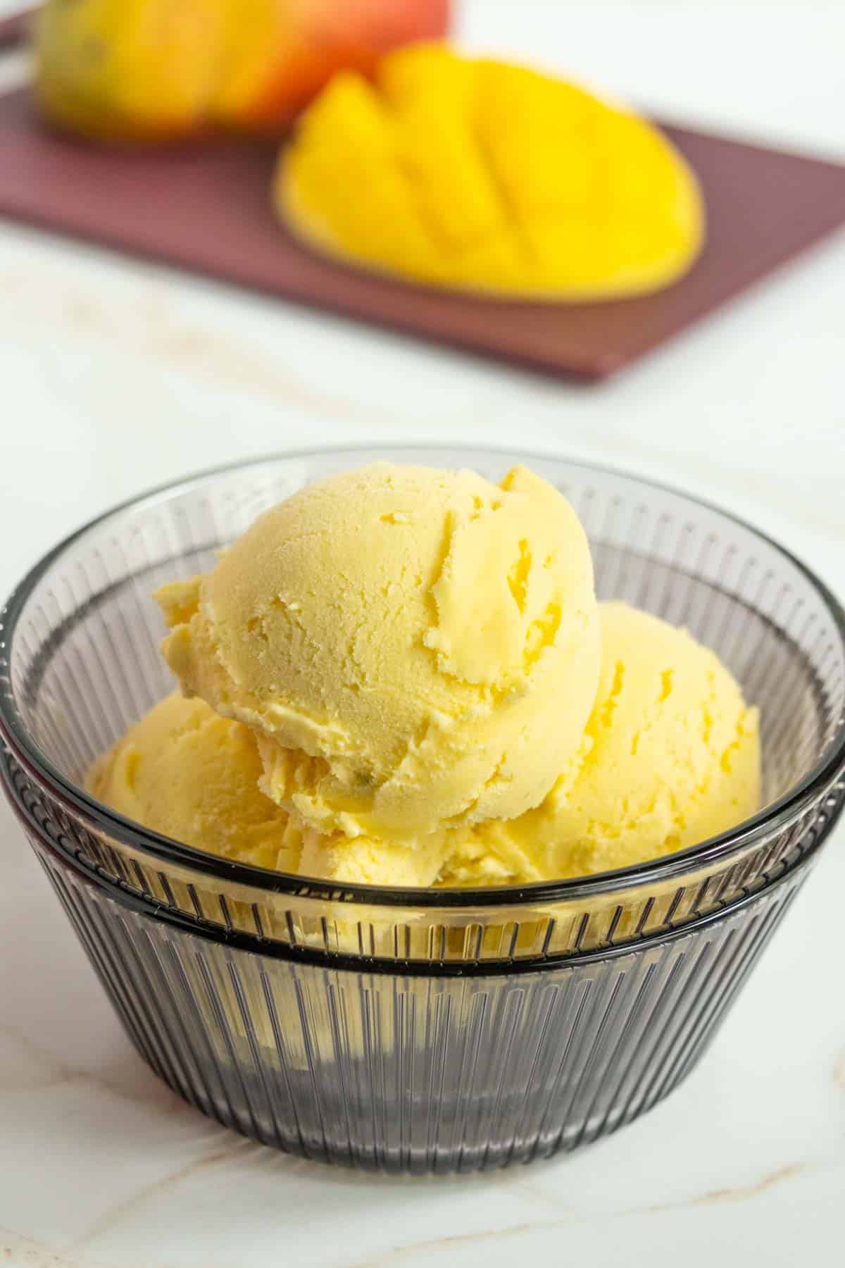 A glass bowl filled with three scoops of yellow mango ice cream, with fresh mango pieces on a cutting board in the background.