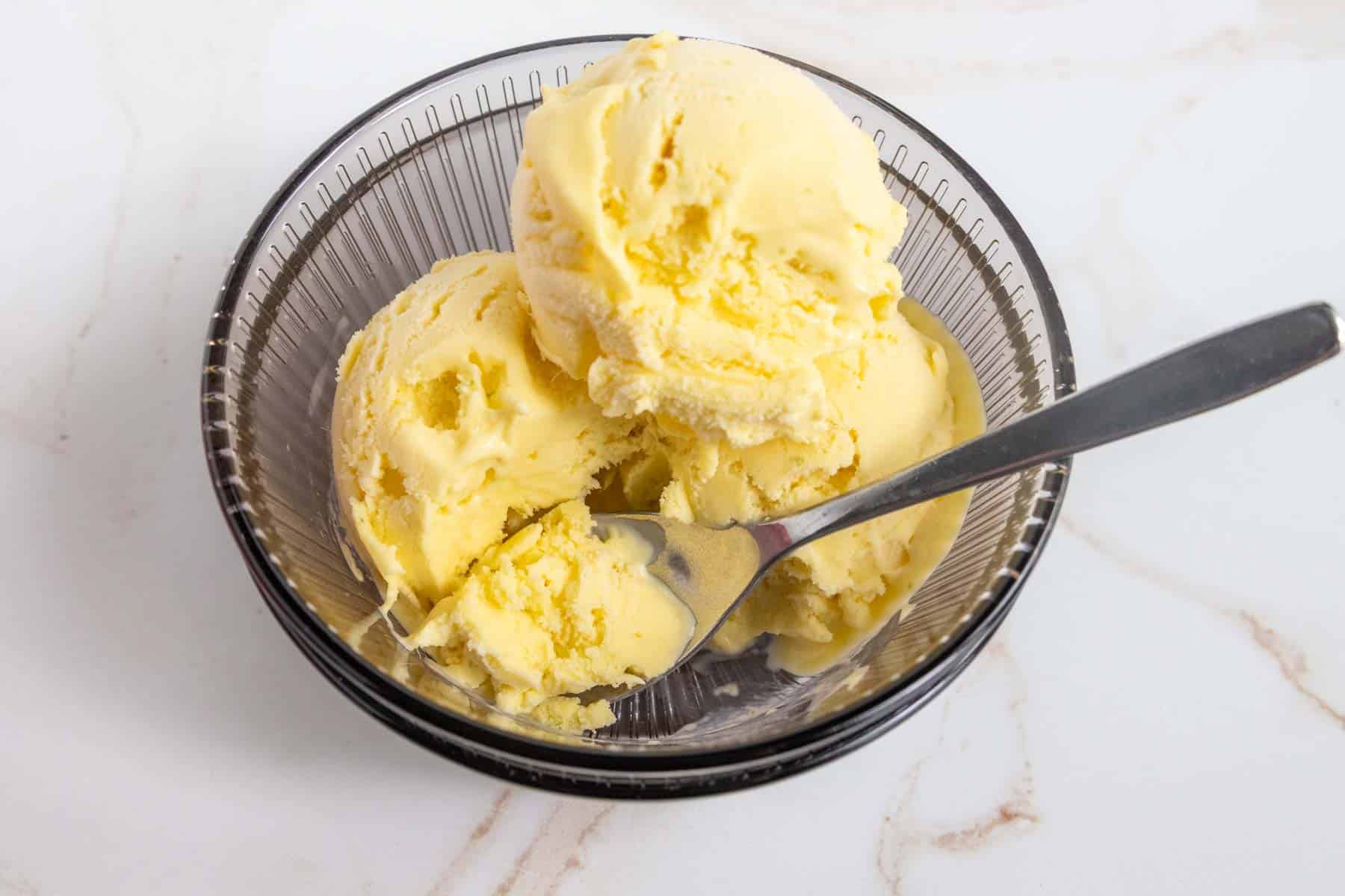 A clear glass bowl with three scoops of yellow ice cream and a metal spoon on a white surface.
