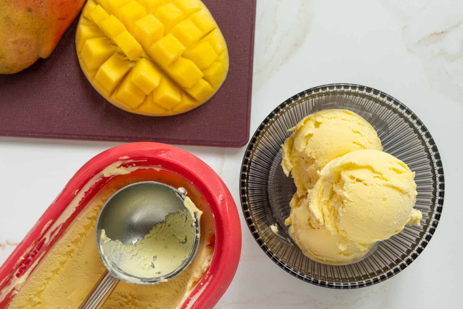 A bowl with scoops of mango ice cream next to a container of ice cream with a scoop, and a sliced mango on a cutting board.