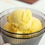 A clear bowl filled with three scoops of yellow mango ice cream, with a plate of more mango ice cream in the background. Text reads "Mango Ice Cream.