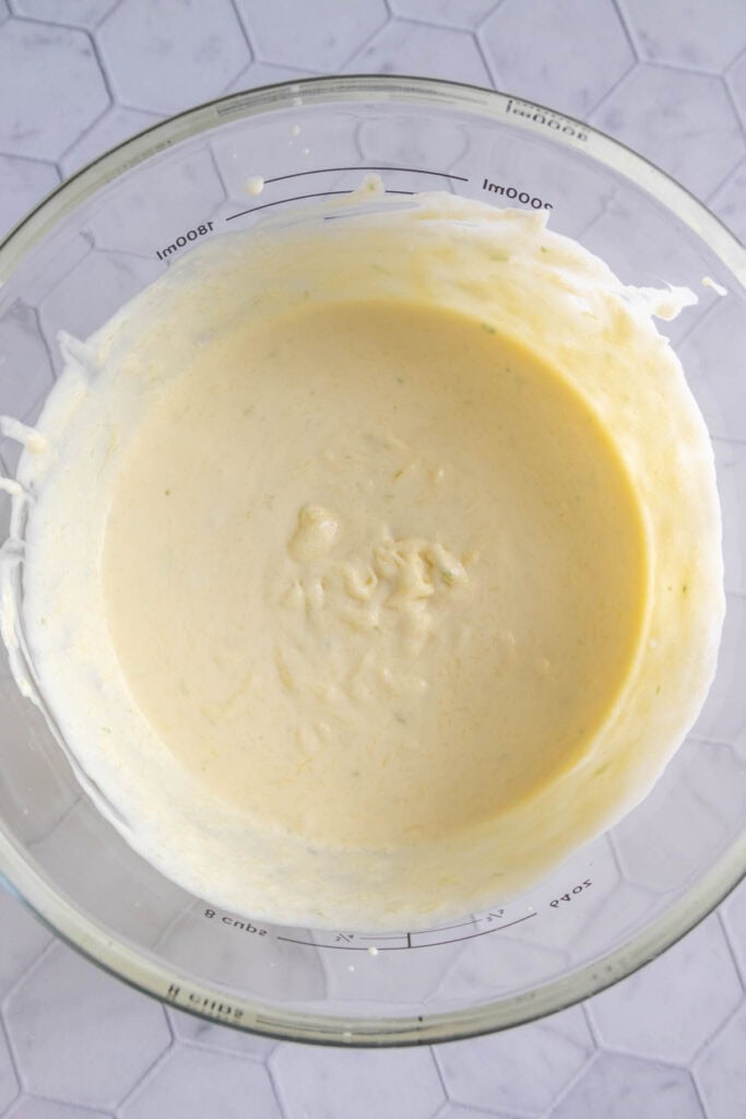 A glass mixing bowl filled with a smooth, pale yellow batter sits on a white hexagonal tile surface.