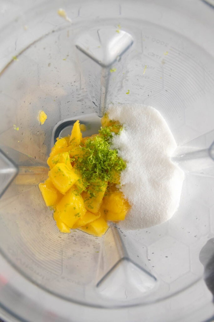Chopped mango, lime zest, and granulated sugar in a blender jar, ready to be blended.