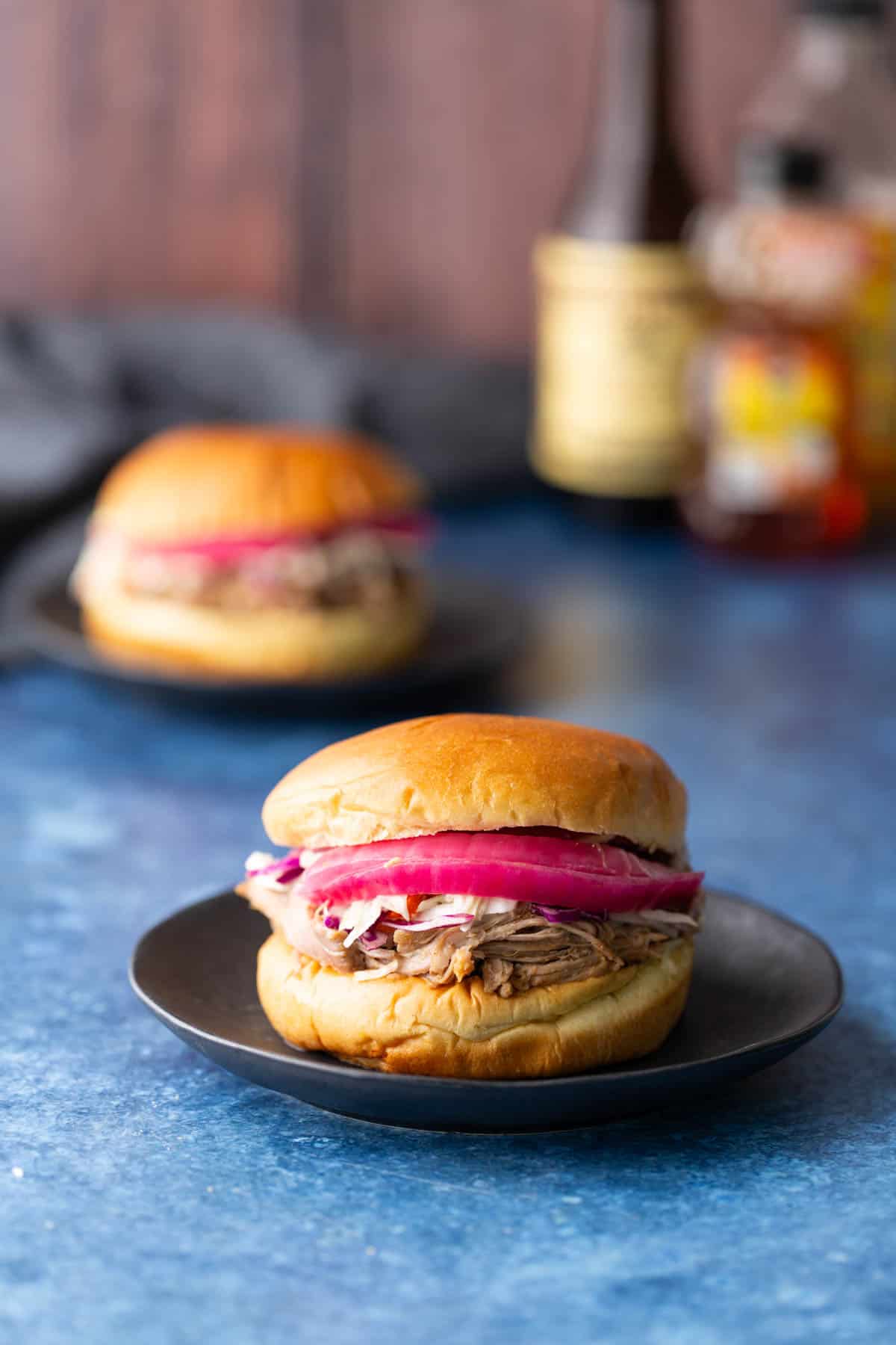 Two pulled pork sandwiches with pickled red onions on black plates, set on a blue surface with blurred bottles and another sandwich in the background.