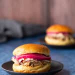 A pulled pork sandwich with pickled onions and coleslaw on a black plate, with a similar sandwich blurred in the background.