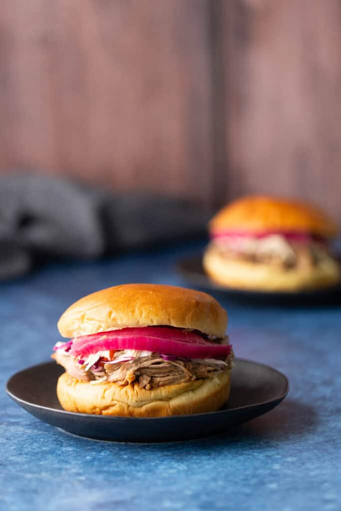 Slow Cooker Pulled Pork A pulled pork sandwich with pickled onions and coleslaw on a black plate, with a similar sandwich blurred in the background.
