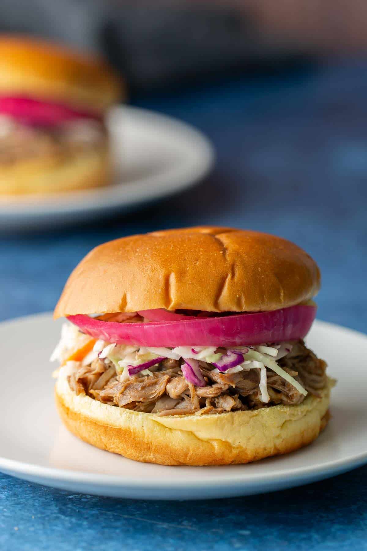 A pulled pork sandwich with coleslaw and sliced red onions on a white plate, with another sandwich blurred in the background.