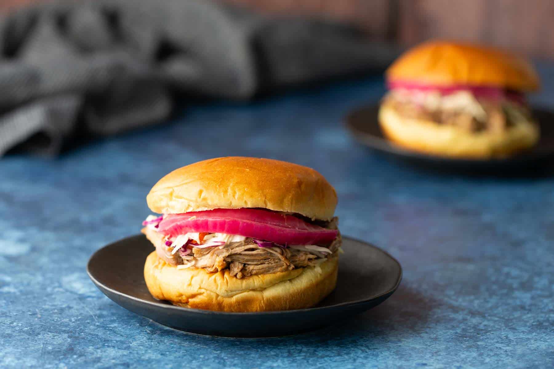 A pulled pork sandwich with pickled onions and slaw sits on a black plate, with another sandwich blurred in the background on a blue surface.