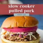 A pulled pork sandwich topped with coleslaw and pickled red onions on a bun, served on a white plate. Text overlay reads "slow cooker pulled pork.
